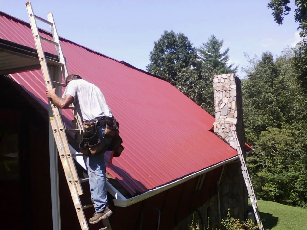 Roofer on a ladder beside a red metal roof and stone chimney — Soffit & Fascia Repair by Roofing Brothers in Scottdale